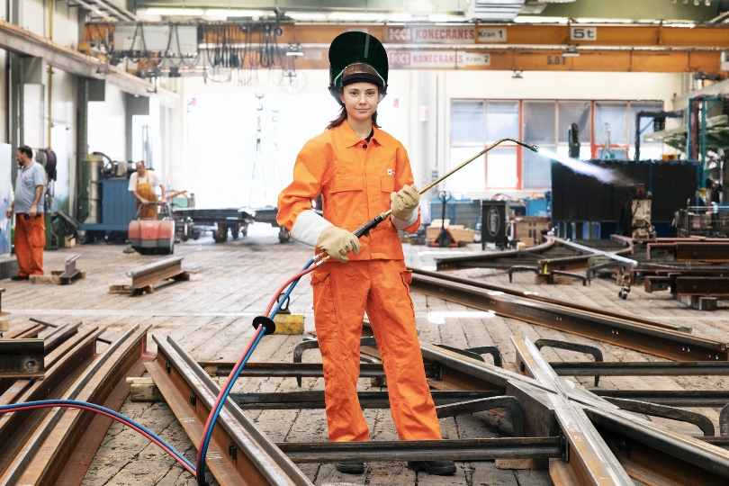 Dame mit Visierhelm hält ein Schweißgerät und steht in einer Halle der Wiener Stadtwerke zwischen Gleisen